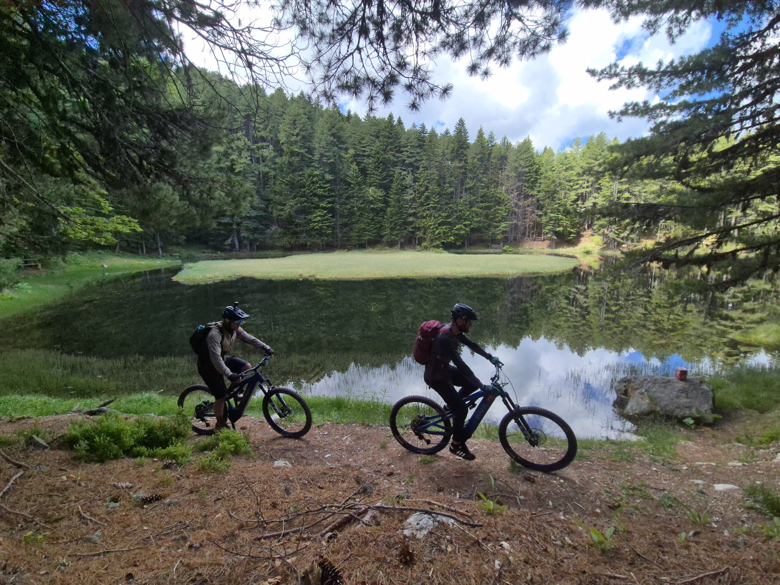Two riders at the shore of Visitor Lake, ringed by pine forest